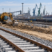 AI-generated illustration of railway construction with an excavator, new tracks and industrial port infrastructure; not a photograph of the Port of Police or the actual contaminated section
