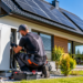 Technician servicing a residential heat pump beside a Polish home with rooftop solar panels